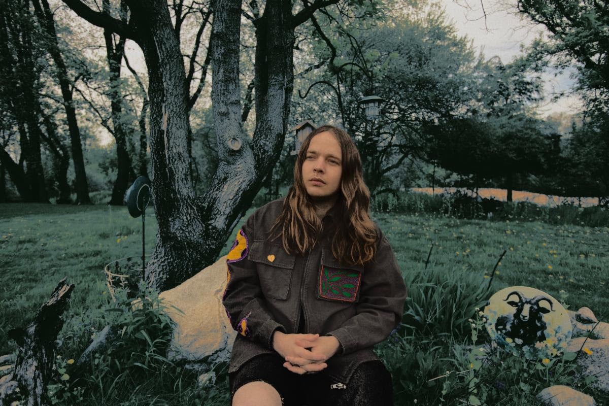 A picture of Billy Strings sitting down in a grassland.